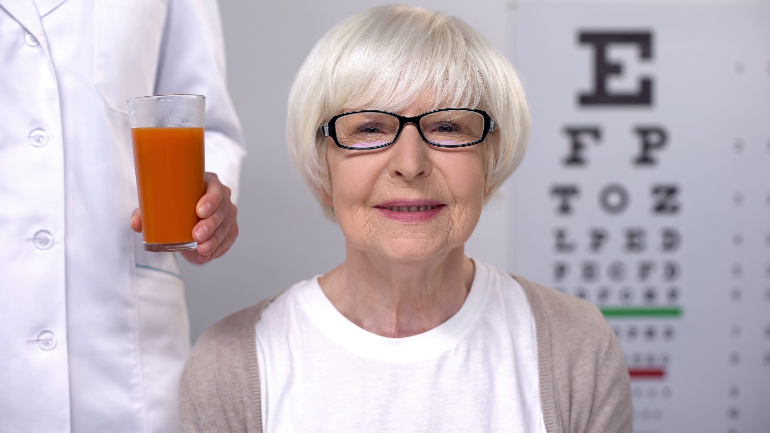 Optician giving fresh carrot juice to happy aged lady, maintaining good vision